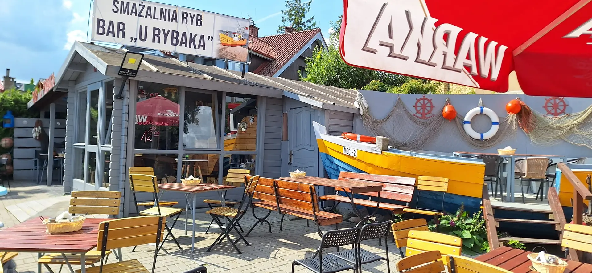 Ogródek letni Baru U Rybaka w Mechelinkach z drewnianymi ławami i kolorową łodzią rybacką